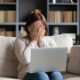 Woman on sofa with her hands over her eyes and a computer on her lap.